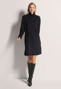 Schwarzes Strickkleid mit hochgeschlossenem Kragen, langen Ärmeln und seitlichen Reißverschlusstaschen, kombiniert mit kniehohen schwarzen Stiefeln. Glatte Textur und taillierte Passform.