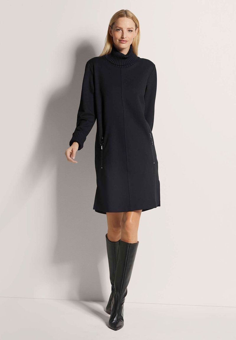 Schwarzes Strickkleid mit hochgeschlossenem Kragen, langen Ärmeln und seitlichen Reißverschlusstaschen, kombiniert mit kniehohen schwarzen Stiefeln. Glatte Textur und taillierte Passform.