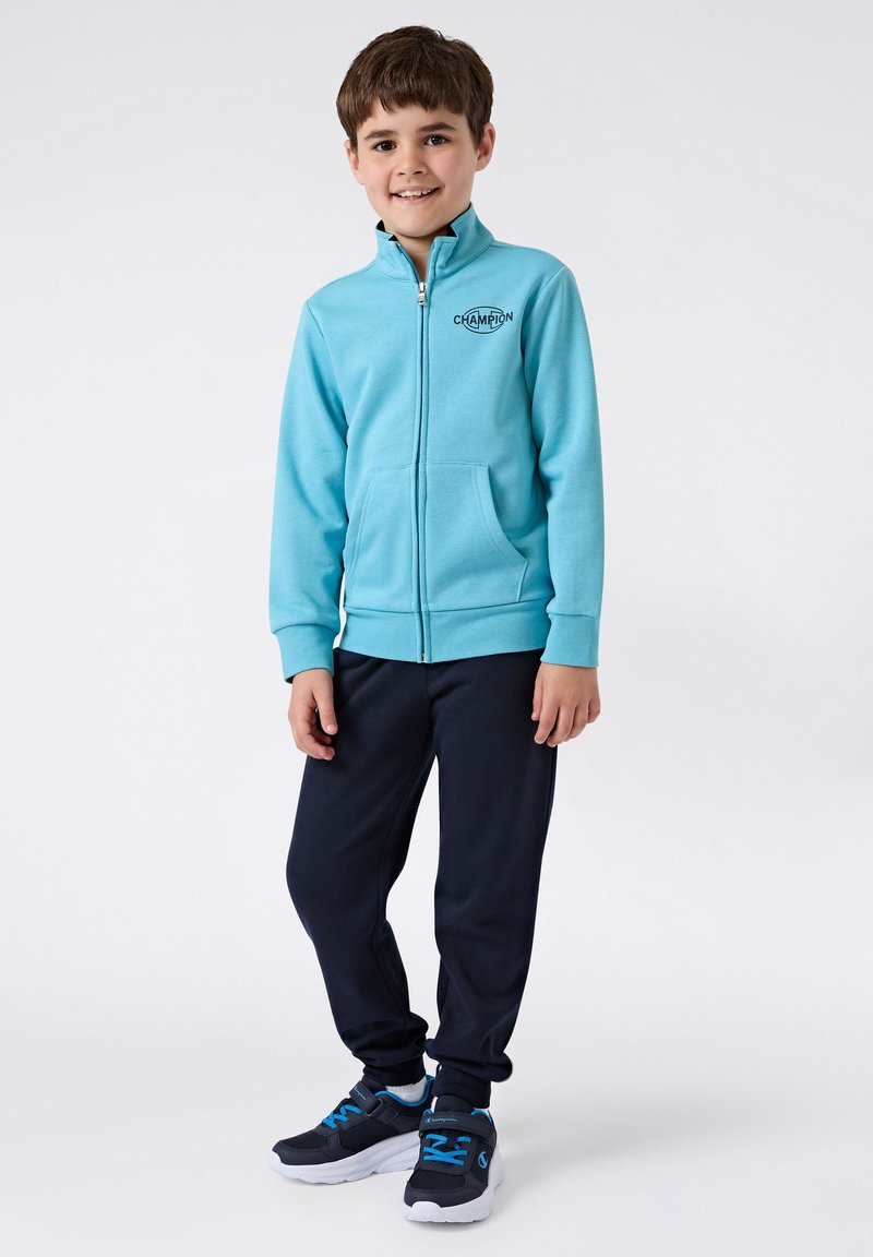 Felpa con zip azzurra chiara con tasche frontali, pantaloni della tuta blu navy e sneakers nere con accenti blu. Abbigliamento sportivo giovanile semplice.