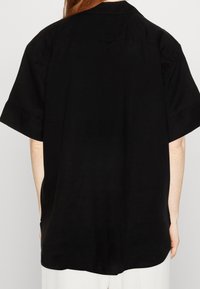 Chemise noire à manches courtes avec une coupe décontractée, en matière texturée, présentant une couture au dos et un ourlet arrondi.