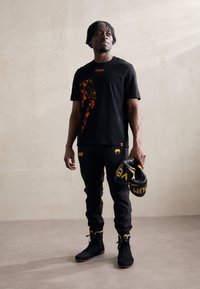 Sort t-shirt med et orange camo-design, parret med sorte sweatpants og high-top sko. Sorte boksehandsker med guldskrift i hånden.