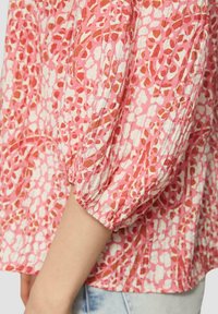 Rosa gemusterte Bluse mit Puffärmeln, die unregelmäßige Formen in Rot und Weiß aufweist, gefertigt aus einem strukturierten Stoff.