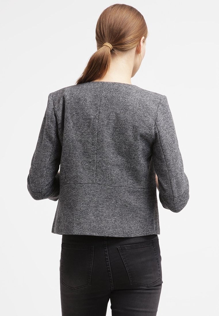 Blazer grigio a textura con una silhouette aderente, maniche lunghe e una cucitura posteriore pulita. Abbinato a pantaloni neri aderenti.