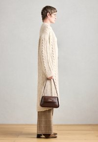 Cardigan crema a maglia con motivi a treccia, borsa marrone, pantaloni a quadri marroni e stivali marroni con punta. Vista laterale, sfondo minimal.