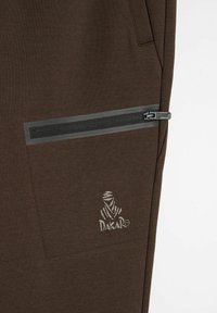 Brązowe dresowe spodnie z boczną kieszenią zamykaną na czarny zamek błyskawiczny z szarym paskiem ozdobnym, z białym logo "DAKAR".