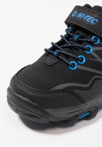 Schwarzer Wanderschuh mit einem strukturierten Obermaterial aus Stoff, blauen Akzenten und reflektierendem grauen Design. Verfügt über Schnürsenkel und einen Klettverschluss.