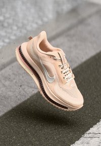 Tenis Nike roz deschis pentru alergare, cu talpă cu aer comprimat, suspendat deasupra unui paviment texturat, cu sireturi legate.