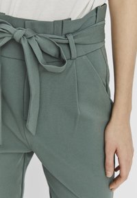 Green high-waisted trousers with a tied belt, featuring pleats and a smooth fabric texture. Close-up view of waistband and hand holding the fabric.