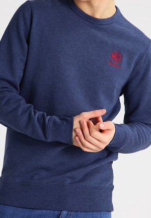 Persona che indossa una felpa Reebok blu navy con logo rosso, con le mani unite in vita, visibile dalle spalle ai fianchi.