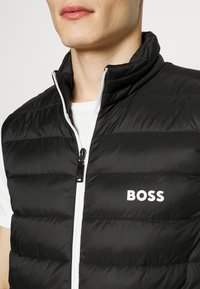 Svart puffväst med horisontell quiltstruktur, vit dragkedja och "BOSS"-logotyp tryckt i vitt på vänster sida.