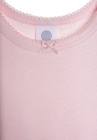 Top in cotone rosa chiaro con texture a coste, delicata rifinitura a scalloped e un piccolo fiocco accentato vicino al collo, con etichetta interna.