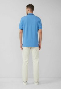 Hellblaues Polohemd mit kurzen Ärmeln und Kragen, kombiniert mit cremefarbener Hose, vor einem neutralen Hintergrund.