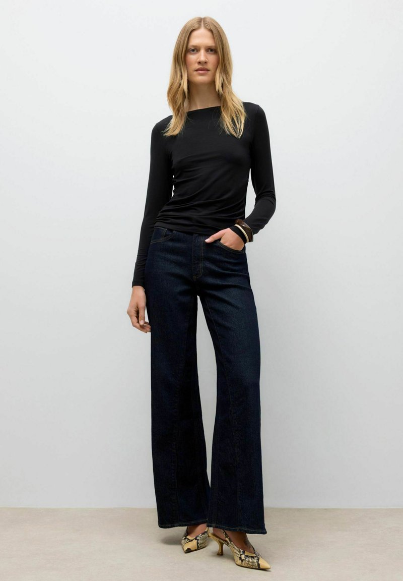 Zwarte top met lange mouwen, gecombineerd met hooggewaisted, wijde jeans van donker denim en gepatternde hakken met een geel en zwart kleurenschema.