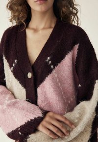 Adīta džemperis ar trīsstūra rakstu, kas ietver bordo, rozā un krēmdzeltenas krāsas. Iekļauta pogu aizdare un teksturēta auduma sastāvs.