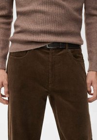 Pantalones de pana marrones con corte recto y canalones verticales, con bolsillos frontales y un cinturón negro. La tela texturizada añade profundidad.