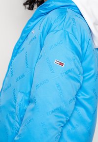 Giacca a vento blu con un motivo ripetitivo del logo "TOMMY JEANS". Include un piccolo emblema del marchio e un cappuccio. Il materiale appare liscio e lucido.