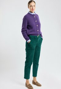 Cardigan viola lavorato a maglia con bottoni in legno, abbinato a pantaloni in corduroy verde scuro. Scarpe marrone maculate completano l'outfit.