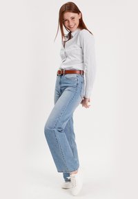 Jean droit bleu clair taille haute, associé à une chemise blanche boutonnée et une ceinture marron ; modèle portant des baskets blanches.