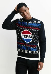 Schwarzer Strickpullover mit großem Pepsi-Logo, blauen und roten Akzenten sowie Schneeflockenmuster mit dem Text "Es ist die Saison, spritzig zu sein."
