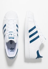 White leather sneakers with three navy blue stripes, textured toe cap, and white laces. Features a padded collar and branded tongue.