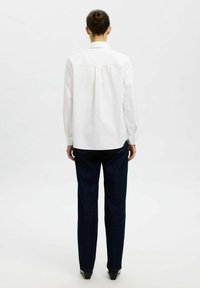 Camisa blanca de manga larga con un pliegue en la parte posterior, combinada con jeans de pierna recta en azul oscuro. El conjunto presenta elementos de diseño minimalistas.
