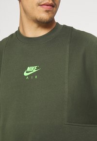 Olive green Nike sweatshirt featuring front pocket, ribbed neck, and a glowing green logo with "AIR" text. Soft, textured fabric.