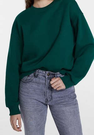 Persoon draagt een donkergroene lange mouwen trui, ingestopt in lichtgrijze jeans met hoge taille en een knoop- en ritssluiting.