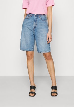 Person iført blå cowboyshorts til knæet, en pink kortærmet shirt og sorte sandaler med åbne tæer, står foran en ensfarvet baggrund.