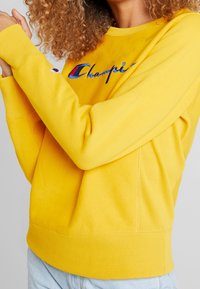 Felpa gialla con logo "Champion" ricamato in rosso e blu. Presenta un colletto e polsini a costine, realizzata in morbido cotone.