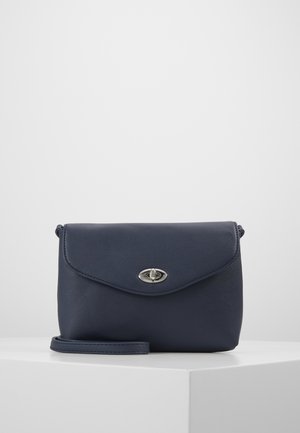 Sac bandoulière en cuir bleu marine avec rabat et fermoir à torsion argenté, reposant sur une surface blanche sur un fond gris clair.
