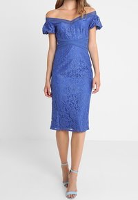 Modré krajkové šaty odhalené na ramena s přiléhavým střihem, texturovaným květinovým vzorem a krátkými nafouklými rukávy, doplněné světlomodrými sandály na podpatku.