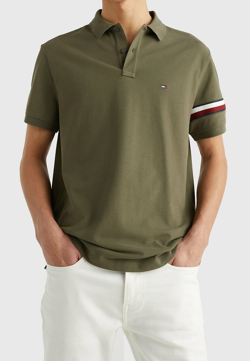 Homme portant un polo vert olive avec des rayures rouges, blanches et bleu marine sur les bras, associé à un pantalon blanc, les mains dans les poches, sur un fond neutre.