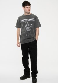 Gray cotton t-shirt featuring a white graphic print of a hand with the text "MON THE BIFF" above. Paired with black trousers and boots.