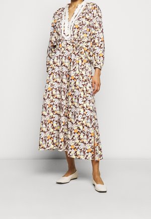 Kobieta ubrana w długą sukienkę z długim rękawem i dekoltem w serek, w kwiatowy wzór w kolorach brązu, pomarańczu i bieli, do tego białe płaskie buty.