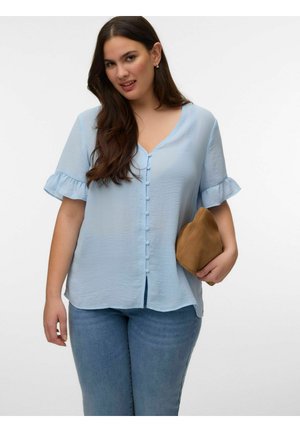 Vrouw met lang bruin haar, gekleed in een lichtblauwe blouse met knopen en gerimpelde mouwen, en een spijkerbroek, houdend een bruine clutch.