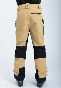 Beige og navy skibukser med en tætsiddende design, lynlåslommer og justerbar talje. Lavet af vandafvisende materiale til udendørs brug.