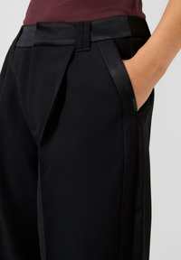 Osoba w czarnych spodniach z satynowym wykończeniem przy kieszeniach i pasku, ręka swobodnie umieszczona w prawej kieszeni.