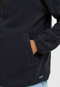 Mørk marineblå fleece-pullover med halv glidelås i halsen, frontlomme og elastiske mansjetter. Har teksturert stoff og et lite merkelapp nederst.