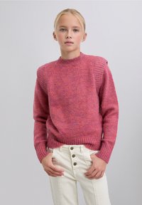 Suéter rosa de punto con cuello redondo, detalles texturizados en los hombros, puños y dobladillo acanalados. Llevado sobre pantalones blancos de talle alto con botones.