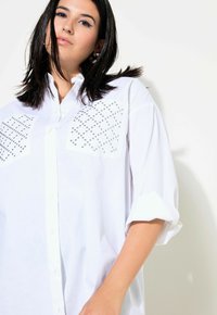 Camicia bianca oversize con bottoni, caratterizzata da due grandi tasche decorate con borchie argentate. Le maniche arrotolate aggiungono un tocco di struttura.