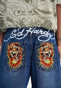 Denum-shortsit sinim pesuga, millel on tikandid kolju kujutistega, mis on kaunistatud erksate oranžide ja kollaste aktsentidega tagakottidel. "Ed Hardy" logo valges kirjas.