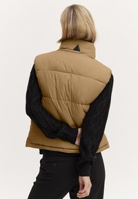 Wattierte Weste aus beigem Stoff mit hohem Kragen, ausgestattet mit einem schwarzen Strickärmel mit einem Kabelmuster, glatter Textur und dreieckigem Akzent.