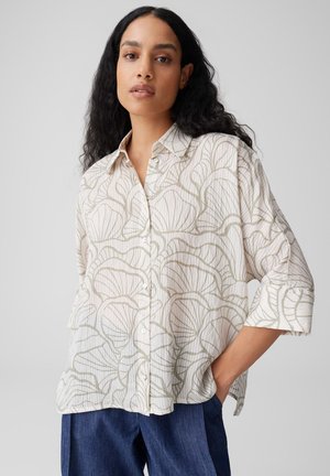 Femme aux longs cheveux noirs portant une chemise blanche ample à motifs et un pantalon bleu foncé, debout avec une main dans la poche.