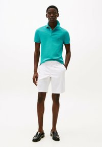 Türkises Poloshirt mit Kragen, kombiniert mit weißen Shorts und schwarzen Lederloafers. Glatte Textur, taillierte Passform.