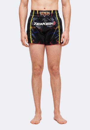 Hombre vistiendo pantalones cortos negros de la marca Venum Tekken 8 con acentos amarillos y rojos, de pie descalzo sobre un fondo liso.