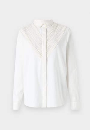 Chemise blanche avec un col boutonné, présentant un détail en crochet décoratif au niveau du décolleté en V. Manches longues et texture lisse.