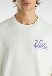 Camiseta corta de manga corta en color crema claro con cuello redondo. Presenta un logo de Vans en color púrpura impreso en el pecho. Confeccionada en suave material de algodón.