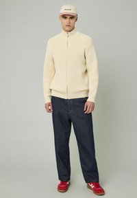 Pull zippé beige à rayures, jean bleu foncé ample, baskets rouges et casquette beige avec texte. Design simple et tissu texturé.