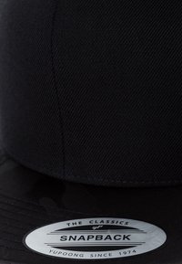 Fekete snapback sapka textúrált anyaggal, lapos erővel és kör alakú címkével, amely tartalmazza a márka és a modell részleteit.