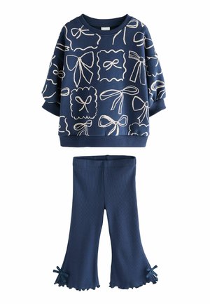 Marineblaues Sweatshirt mit weißen Schleifenmotiven und gerippten Bündchen, kombiniert mit passenden ausgestellt geschnittenen Hosen mit Rüschensaum und Schleifen an den Seiten.